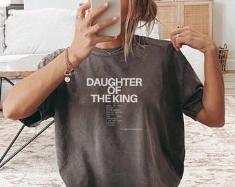 May include: A charcoal grey short-sleeve t-shirt featuring the white text DAUGHTER OF THE KING in a bold sans-serif font, followed by smaller printed scripture text from 2 Corinthians 5:19, with a crew neckline and a relaxed fit design.