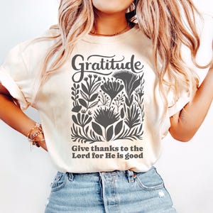 May include: A cream-colored short-sleeve t-shirt featuring a dark gray graphic print. The design includes the word Gratitude in a script font above a collection of stylized floral illustrations and the text Give thanks to the Lord for He is good.
