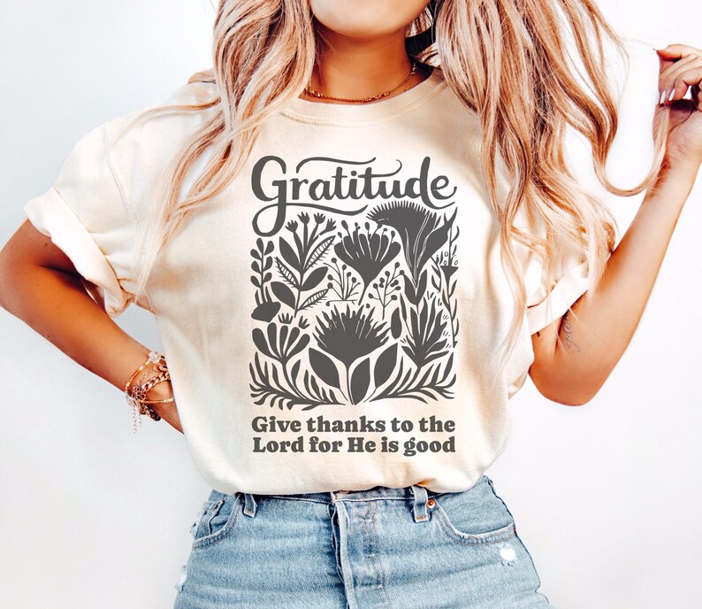 May include: A cream-colored short-sleeve t-shirt featuring a dark gray graphic print. The design includes the word Gratitude in a script font above a collection of stylized floral illustrations and the text Give thanks to the Lord for He is good.