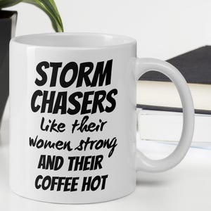 Könnte beinhalten: Eine weiße Keramiktasse mit schwarzem Text, der "Storm Chasers like their women strong and their coffee hot" lautet.