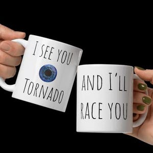 Könnte beinhalten: Zwei weiße Keramikbecher mit schwarzem Text. Der Becher links sagt "I see you tornado" mit einer blauen Augengrafik. Der Becher rechts sagt "And I'll race you".