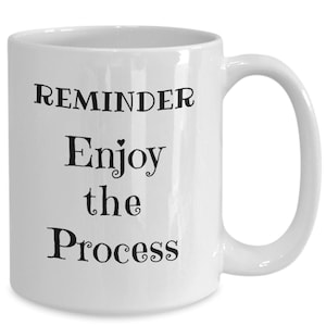 May include: White ceramic coffee mug with black text that reads "REMINDER Enjoy the Process".