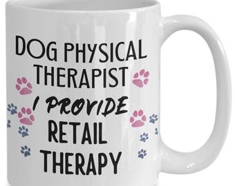 Pode incluir: Caneca de café de cerâmica branca com texto preto que diz "Dog Physical Therapist I Provide Retail Therapy". O texto é cercado por pegadas de patas rosa e azuis.