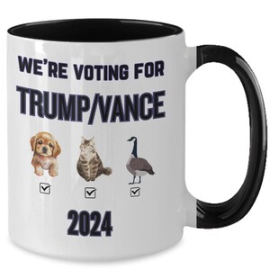 May include: White ceramic coffee mug with a black rim and handle. The mug has a graphic of a dog, cat, and goose with checkboxes next to each. The text on the mug reads "We're voting for Trump/Vance 2024".