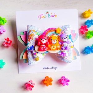 May include: A hair bow featuring a white glittery base with a Care Bears pattern, topped with three miniature Care Bear figurines in red, orange, and purple, a rainbow, and a yellow star. The bow is attached to a white card labeled Tini Bows.