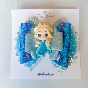 May include: A multi-layered hair bow featuring blue and silver glitter fabric with a printed pattern of a blonde character in a blue dress. A central 3D resin figure of the same character is attached. The display card reads Tini Bows and tinibowsdesign.