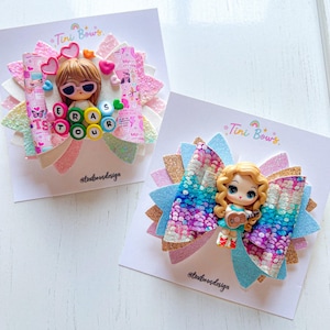 May include: Two hair bows on display cards. The left bow features a doll with sunglasses and beads spelling ERAS TOUR. The right bow features a doll with a guitar. Both bows are made of multi-coloured glittery fabric with layered petal shapes.