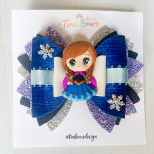 May include: A multi-layered hair bow featuring a central cartoon character figurine with brown hair and a blue dress. The bow is constructed from blue velvet, silver glitter, and purple glitter fabric, accented with two silver snowflake charms and light blue ribbon.