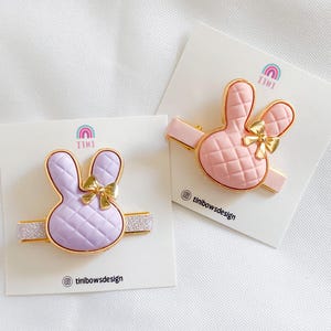 May include: Two pink and purple bunny hair clips with gold bows and glitter accents. The clips are on white cards with the text "Tini" and "tinibowsdesign".