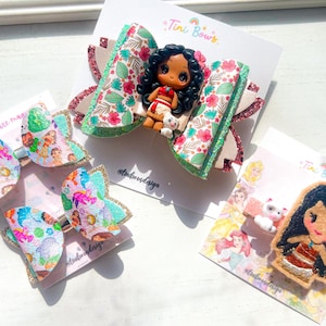 May include: Three hair accessories on display cards labeled Tini Bows. One large bow features a Moana character figurine with floral patterns and glittery edges. Another bow has a colorful Moana print. A felt hair clip depicts a cartoon character.
