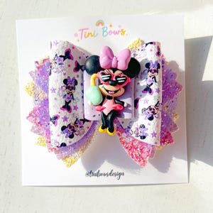 May include: A multi-layered hair bow featuring a white faux leather center with a repeating Minnie Mouse print, accented by layers of purple and pink glitter fabric. A 3D resin charm of Minnie Mouse wearing heart-shaped sunglasses is attached.