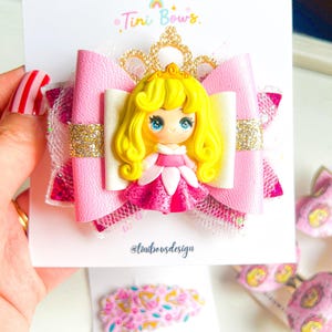 May include: A hair bow featuring a central figurine of a blonde princess in a pink dress, layered with pink faux leather, white glitter accents, and a gold crown cutout. The display card includes the text Tini Bows and @tinibowsdesign.