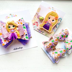 May include: Three hair accessories on display cards labeled Tini Bows. One large purple and gold glitter bow features a blonde character figurine. A second card holds a felt character patch. A third card displays two smaller bows with a character print.
