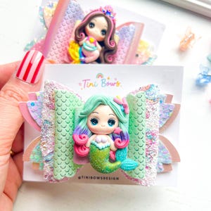 May include: A hair bow featuring a central 3D mermaid figurine with mint green and purple hair, a glittery green tail, and a seashell top. The bow is constructed from layered materials including mint green scale-patterned fabric and glittery accents. Text reads Tini Bows.