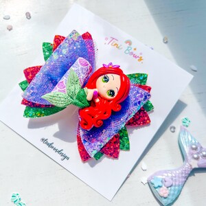 May include: A hair bow featuring layers of purple, red, and green glitter fabric with a central figurine of a red-haired mermaid with a green tail. The bow is attached to a white display card printed with the text Tini Bows and tinibowsdesign.