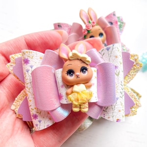 May include: A hair bow featuring a central 3D figurine of a rabbit wearing a yellow tutu and headband. The bow is constructed from layers of lavender faux leather, white material with a floral print, and gold glitter accents on the outer edges.