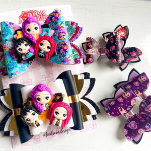 Pode incluir: Quatro laços de cabelo com figuras estilo chibi de cabelo rosa, preto e vermelho. Os laços exibem padrões de notas musicais, estrelas e o texto "HUNTRAX". Os designs incorporam texturas de couro sintético brilhante, dourado metálico e preto.