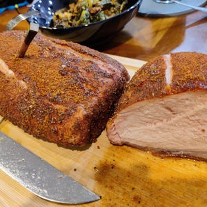 May include: A large, seasoned roast, with a meat thermometer inserted, sits on a wooden cutting board. A portion of the roast has been sliced, revealing the cooked meat. A large knife is placed on the board next to the roast. The roast is a reddish-brown color.