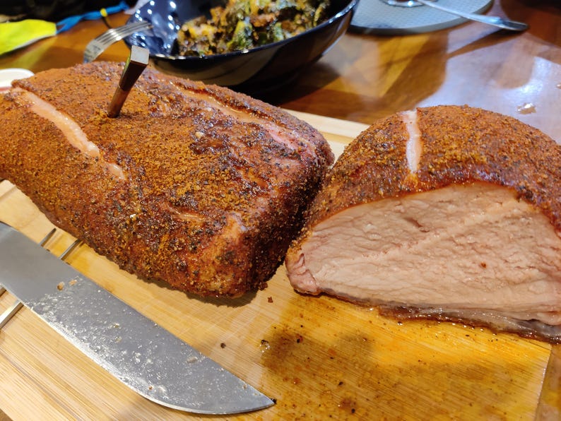 May include: A large, seasoned roast, with a meat thermometer inserted, sits on a wooden cutting board. A portion of the roast has been sliced, revealing the cooked meat. A large knife is placed on the board next to the roast. The roast is a reddish-brown color.