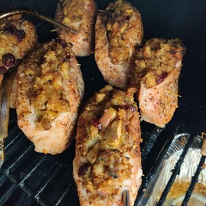 May include: Close-up of several cooked chicken breasts stuffed with a breadcrumb mixture, apples, and cranberries. The chicken is on a black grill, with a metal fork and tongs visible. The dish appears to be seasoned and ready to eat.
