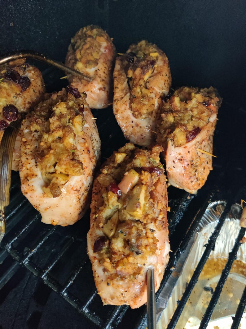 May include: Close-up of several cooked chicken breasts stuffed with a breadcrumb mixture, apples, and cranberries. The chicken is on a black grill, with a metal fork and tongs visible. The dish appears to be seasoned and ready to eat.