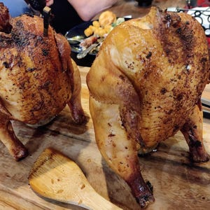 May include: Two roasted chickens with golden-brown skin sit on a wooden cutting board. The chickens are seasoned and appear juicy. A wooden spatula rests near the chickens, suggesting they are ready to be served. A delicious meal.