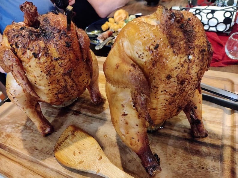 May include: Two roasted chickens with golden-brown skin sit on a wooden cutting board. The chickens are seasoned and appear juicy. A wooden spatula rests near the chickens, suggesting they are ready to be served. A delicious meal.