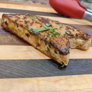 May include: A cooked rectangular piece of food, possibly fish or meat, with a browned exterior and sprinkled with green herbs. It rests on a wooden cutting board with alternating light and dark wood grain. A red and silver kitchen utensil is in the background.