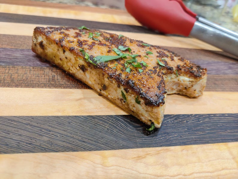 May include: A cooked rectangular piece of food, possibly fish or meat, with a browned exterior and sprinkled with green herbs. It rests on a wooden cutting board with alternating light and dark wood grain. A red and silver kitchen utensil is in the background.