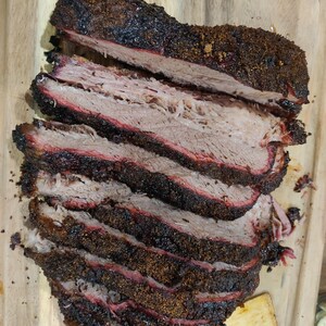 May include: Sliced smoked brisket on a wooden cutting board. The meat has a dark, crusty bark and a pink smoke ring, indicating it was slow-cooked. The brisket is cut into thick slices, showcasing the tender, juicy interior.