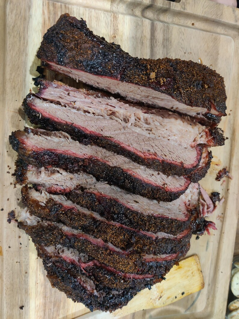 May include: Sliced smoked brisket on a wooden cutting board. The meat has a dark, crusty bark and a pink smoke ring, indicating it was slow-cooked. The brisket is cut into thick slices, showcasing the tender, juicy interior.