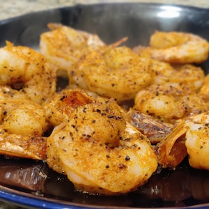 May include: Close-up of cooked shrimp seasoned with spices, arranged on a dark plate. The shrimp are golden brown with visible seasoning and black pepper. The dish is ready to eat, showcasing a seafood meal.