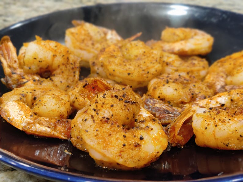 May include: Close-up of cooked shrimp seasoned with spices, arranged on a dark plate. The shrimp are golden brown with visible seasoning and black pepper. The dish is ready to eat, showcasing a seafood meal.
