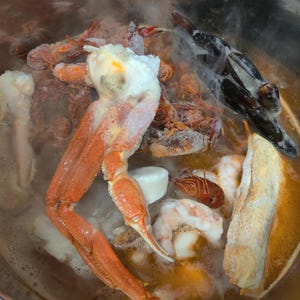 May include: A stainless steel pot filled with seafood, including a large orange crab, shrimp, mussels, and other shellfish. The seafood is partially submerged in a broth, with steam rising from the pot.