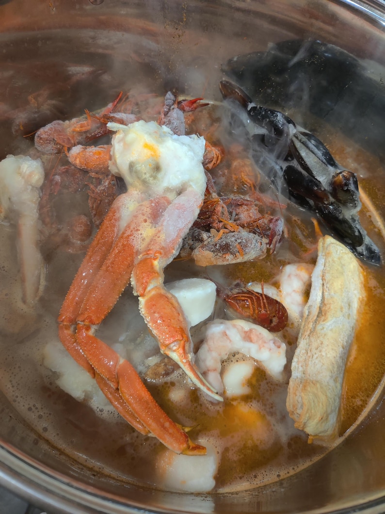 May include: A stainless steel pot filled with seafood, including a large orange crab, shrimp, mussels, and other shellfish. The seafood is partially submerged in a broth, with steam rising from the pot.