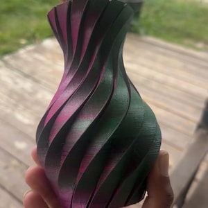 May include: A decorative vase with a unique, swirling design. The vase features alternating bands of green, black, and magenta. The vase is held in a hand and appears to be made of a 3D-printed material. The vase has a wide base and a narrow neck.