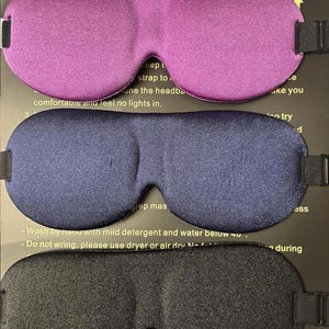 May include: Three sleep masks in purple, navy blue, and black. Each mask has an adjustable strap and is designed to block out light. The masks are made of a soft, smooth material. Instructions are printed on the packaging.