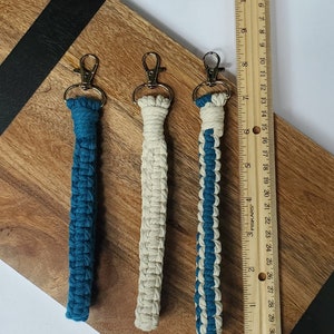 May include: Three keychains with different colour combinations. The keychains are made of woven string in blue and white. The keychains are hanging from a wooden surface.