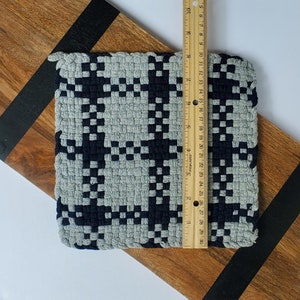 May include: A grey and black woven pot holder with a chequered pattern. The pot holder is measured with a ruler, showing it is approximately 18 cm wide.