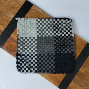 Puede incluir: Un posavasos tejido a cuadros en blanco y negro. El posavasos está hecho de una tela gruesa y texturizada, ideal para proteger tus manos de las superficies calientes.