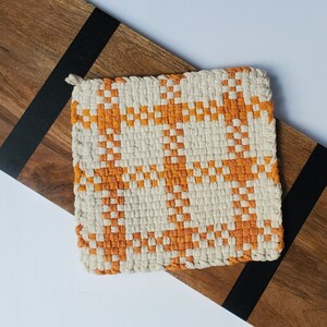 May include: A square, woven potholder with a cream-colored background and an orange geometric pattern. The potholder has a small loop for hanging. It rests on a wooden cutting board with a black stripe.
