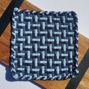 Puede incluir: Un posavasos tejido azul y gris con un patrón geométrico. El posavasos está hecho de un material grueso y duradero y tiene un borde con flecos.