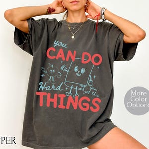 May include: Dark gray t-shirt with the text "You Can Do Hard Things" in red and blue. The shirt features a cartoon apple and book graphic. The shirt is a relaxed fit and has short sleeves.