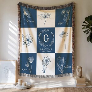Puede incluir: Una manta azul marino y crema con un diseño floral. La manta presenta un patrón de cuadrícula con ilustraciones florales y nombres. El cuadrado central tiene el texto "Grandma Est. 2021" y una letra "G" dentro de una corona.