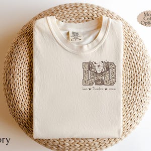 May include: Ivory-colored t-shirt with a crew neck. The shirt features a graphic of the word "DAD" in a camouflage pattern, with a deer head in the center. Below the graphic are the names "Liam", "Theodore", and "emma".