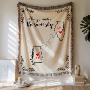 Puede incluir: Manta de color crema con texto e ilustraciones en negro. La manta presenta la frase "Always under the same sky" y contornos de los estados de Alabama e Illinois, conectados por una línea de puntos. Se incluyen los nombres "Sarah" y "Faith".