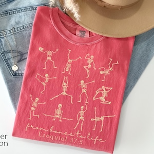 Op de afbeelding: Een watermeloenkleurig T-shirt met een ronde hals met een ontwerp van skeletfiguren in verschillende yogahoudingen. De tekst "from bones to life" en "Ezequiel 37:5" zijn op het shirt gedrukt. Het shirt ligt op een spijkerbroek.