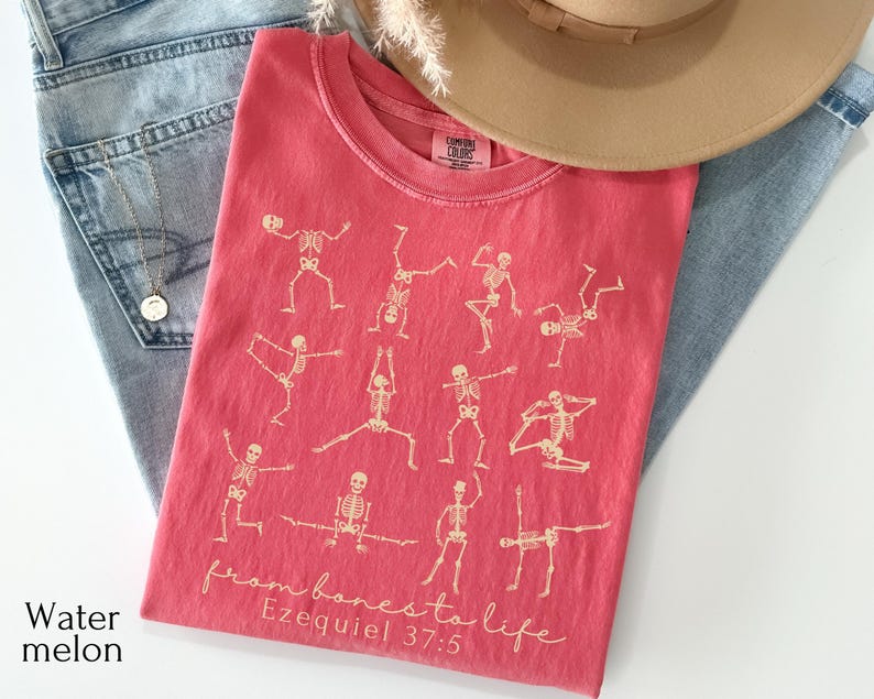 Op de afbeelding: Een watermeloenkleurig T-shirt met een ronde hals met een ontwerp van skeletfiguren in verschillende yogahoudingen. De tekst "from bones to life" en "Ezequiel 37:5" zijn op het shirt gedrukt. Het shirt ligt op een spijkerbroek.