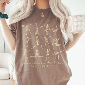 Op de afbeelding: Bruin t-shirt met skeletfiguren in yogahoudingen. De tekst "from bones to life" en "Ezequiel 37:5" is gedrukt. De persoon draagt een mok. Het t-shirt heeft een neutrale kleur.