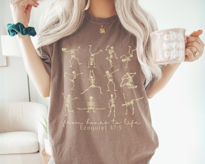 Op de afbeelding: Bruin t-shirt met skeletfiguren in yogahoudingen. De tekst "from bones to life" en "Ezequiel 37:5" is gedrukt. De persoon draagt een mok. Het t-shirt heeft een neutrale kleur.